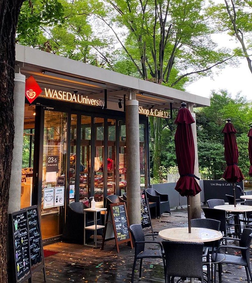 Photo of the Waseda University Uni Shop & Cafe 125. Outdoor seating in the front, trees in the background.