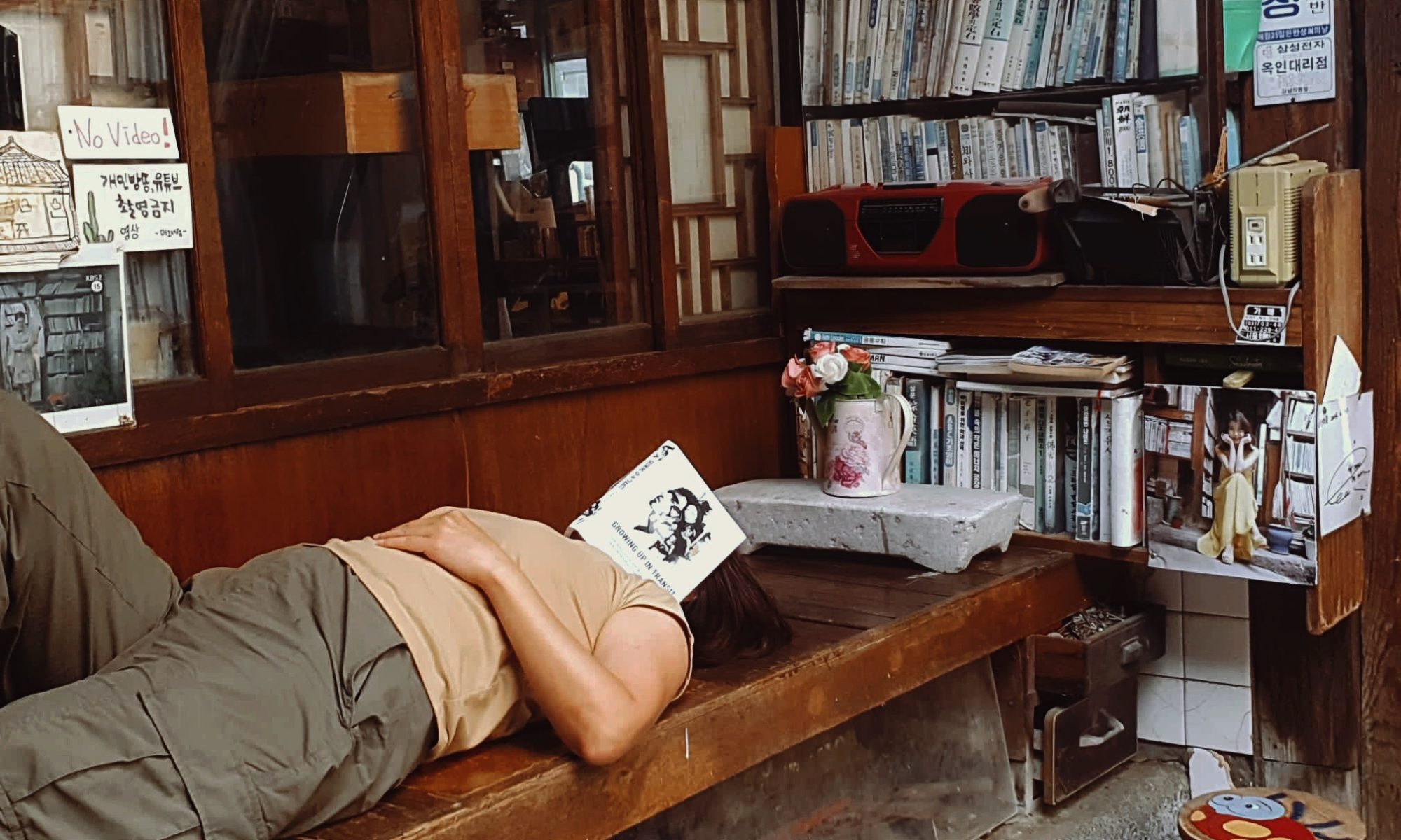 Photo of a woman lying on the engawa of an old Korean bookstore taking a nap. A copy of Growing Up in Transit covering her face. A bookshelf can be seen in the background.